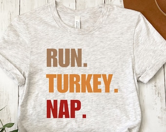 May include: A light gray t-shirt with the words "RUN. TURKEY. NAP." in brown, orange, and red lettering. The shirt is folded and tied at the bottom. A brown hat is visible in the background.