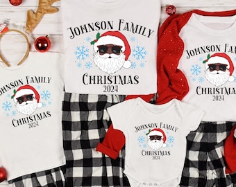 May include: A family Christmas pajama set featuring white tops and a babygrow with a Santa Claus graphic and the text "JOHNSON FAMILY CHRISTMAS 2024". The set includes black and white plaid pyjama bottoms and red accents.