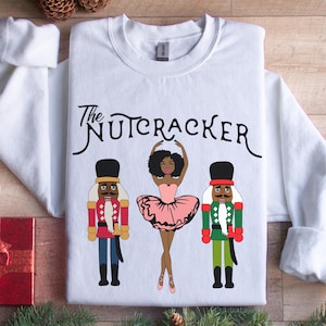 Puede incluir: Sudadera blanca con el texto "The Nutcracker" impreso en negro. El diseño presenta una bailarina negra con un tutú rosa y dos soldados cascanueces, uno en rojo y otro en verde.