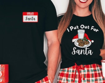 May include: Matching Christmas pajamas set. One person wears a black t-shirt with a name tag that says "Santa". The other wears a black t-shirt with the text "I Put Out For Santa". Both wear red and green plaid pants and hold mugs.