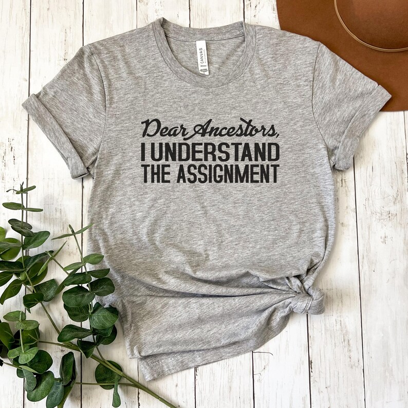May include: Gray t-shirt with the text "Dear Ancestors, I Understand The Assignment" printed in black.