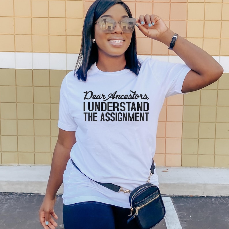 May include: White t-shirt with black text that reads "Dear Ancestors, I Understand The Assignment".