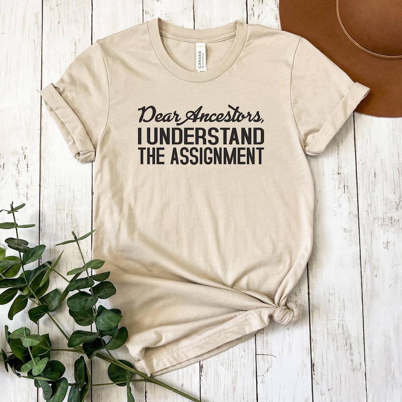 May include: A beige t-shirt with the text "Dear Ancestors, I Understand The Assignment" printed in black.