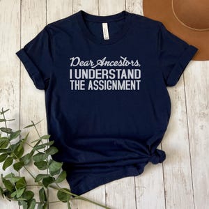 May include: A navy blue t-shirt with the text "Dear Ancestors I Understand The Assignment" printed in white.
