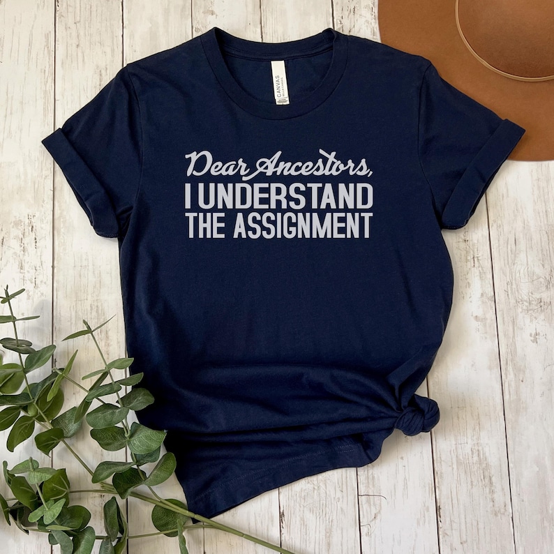May include: A navy blue t-shirt with the text "Dear Ancestors I Understand The Assignment" printed in white.