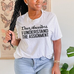 May include: A gray t-shirt with the text "Dear Ancestors, I Understand The Assignment" printed in black.