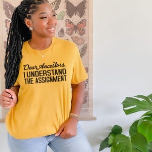 May include: A woman wearing a mustard yellow t-shirt with the text "Dear Ancestors, I Understand The Assignment" printed in black. She is wearing blue jeans and has long black braids.