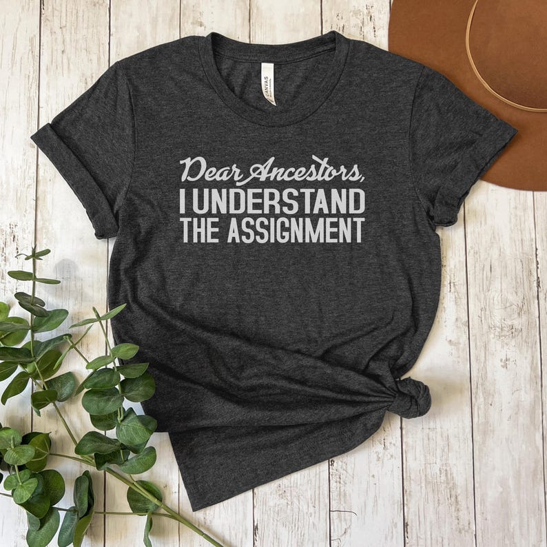 May include: A dark gray t-shirt with the text "Dear Ancestors, I understand the assignment" printed in white.