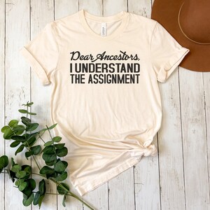 May include: A beige t-shirt with the text "Dear Ancestors, I Understand The Assignment" printed in black.