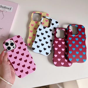Könnte beinhalten: Eine Sammlung farbenfroher Handyhüllen mit Herzmustern. Die Hüllen sind in verschiedenen Farben erhältlich, darunter Rosa, Gelb, Weiß und Burgunderrot, jede mit einem kontrastierenden Herzdesign. Die Hüllen sind für Smartphones konzipiert.
