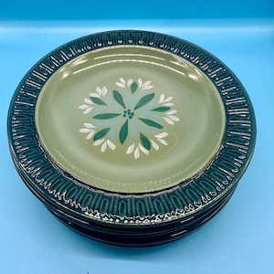 Puede incluir: Una pila de seis platos de cena de cerámica verde y blanca con un diseño floral. Los platos tienen un borde verde oscuro con un patrón texturizado.