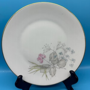 Op de afbeelding: Een wit keramiek bord met een gouden rand en een bloemenontwerp met roze, blauwe en grijze bloemen en bladeren.