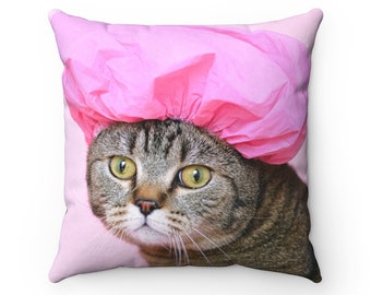 May include: A gray tabby cat wearing a pink shower cap. The cat is looking directly at the camera with a curious expression.
