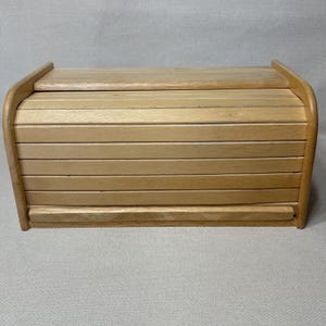 May include: A rectangular wooden bread box featuring a light brown natural wood grain finish. The container has a curved top design with a sliding roll-top door composed of horizontal wooden slats and a solid flat top panel with rounded side edges.