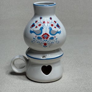 May include: A white ceramic oil warmer featuring a bulbous top section decorated with blue and grey birds, red flowers, and green foliage. The base includes a circular handle and a heart-shaped cutout, with a blue rim accenting the top and base.