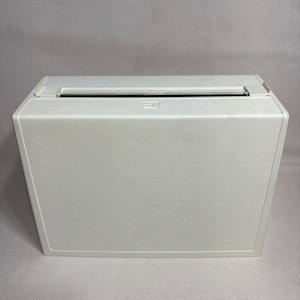 May include: A rectangular off-white plastic storage box with a smooth, matte finish. The box features a wide, horizontal slot opening along the top edge and a small, square-shaped embossed icon centered just below the top opening.