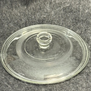 May include: A circular clear glass lid featuring a central knob handle and a decorative concentric ring pattern etched into the surface. The lid has a smooth, raised outer rim and a transparent finish, designed to fit a matching glass container.