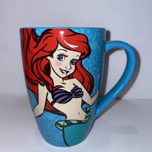 May include: A blue ceramic mug with a colourful illustration of Ariel from The Little Mermaid. The mug has a blue handle and a light blue patterned background. The illustration shows Ariel with red hair and a purple seashell bra.