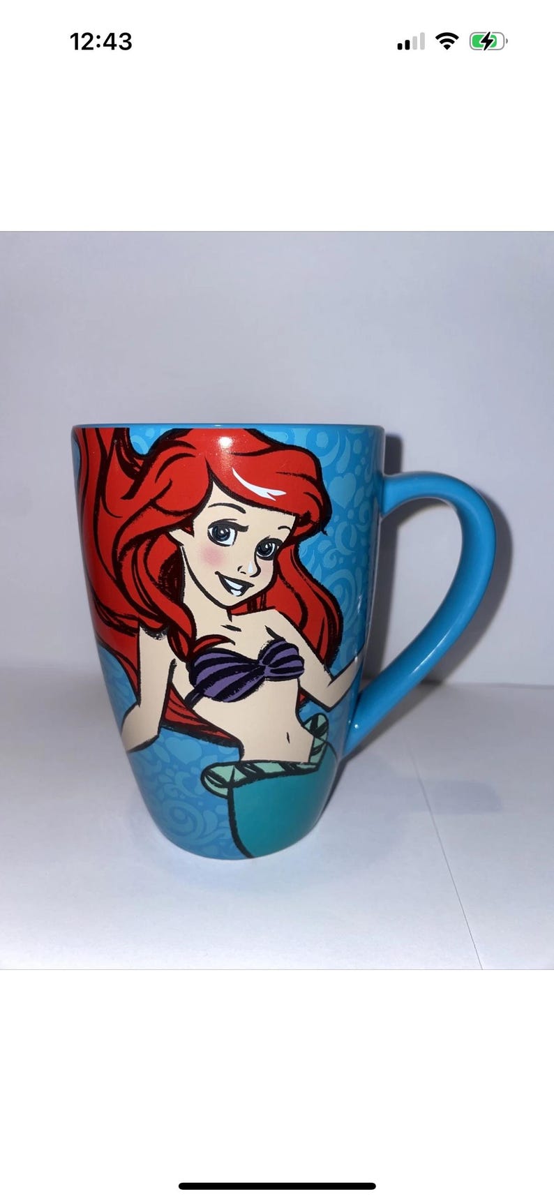 May include: A blue ceramic mug with a colourful illustration of Ariel from The Little Mermaid. The mug has a blue handle and a light blue patterned background. The illustration shows Ariel with red hair and a purple seashell bra.
