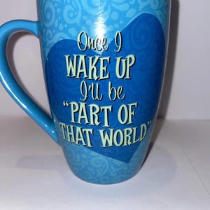May include: A light blue ceramic mug with a handle. The mug features the text "Once I wake up I'll be 'PART OF THAT WORLD'" in white, with a decorative swirl pattern. The mug is a standard size for coffee or tea.