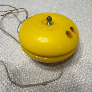 May include: A bright yellow plastic yo-yo featuring a smooth, rounded body with two circular holes on the side and a small metallic center post. A thin, light-colored string is attached and extends outward from the center of the toy.