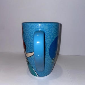 May include: A blue ceramic mug with a curved handle. The mug features a cartoon character design and the text "Thank You". The mug has a light blue handle and a patterned background.