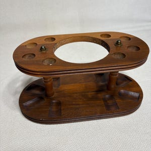 May include: A brown wooden stand with an oval shape, featuring a large central circular opening on the top tier surrounded by eight smaller circular holes. The base tier includes eight recessed slots, and two small metal fasteners are visible.