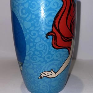 May include: A blue ceramic mug featuring a design of a character with red hair and a white arm. The mug has a light blue background with a pattern of swirls and hearts. The mug is cylindrical.