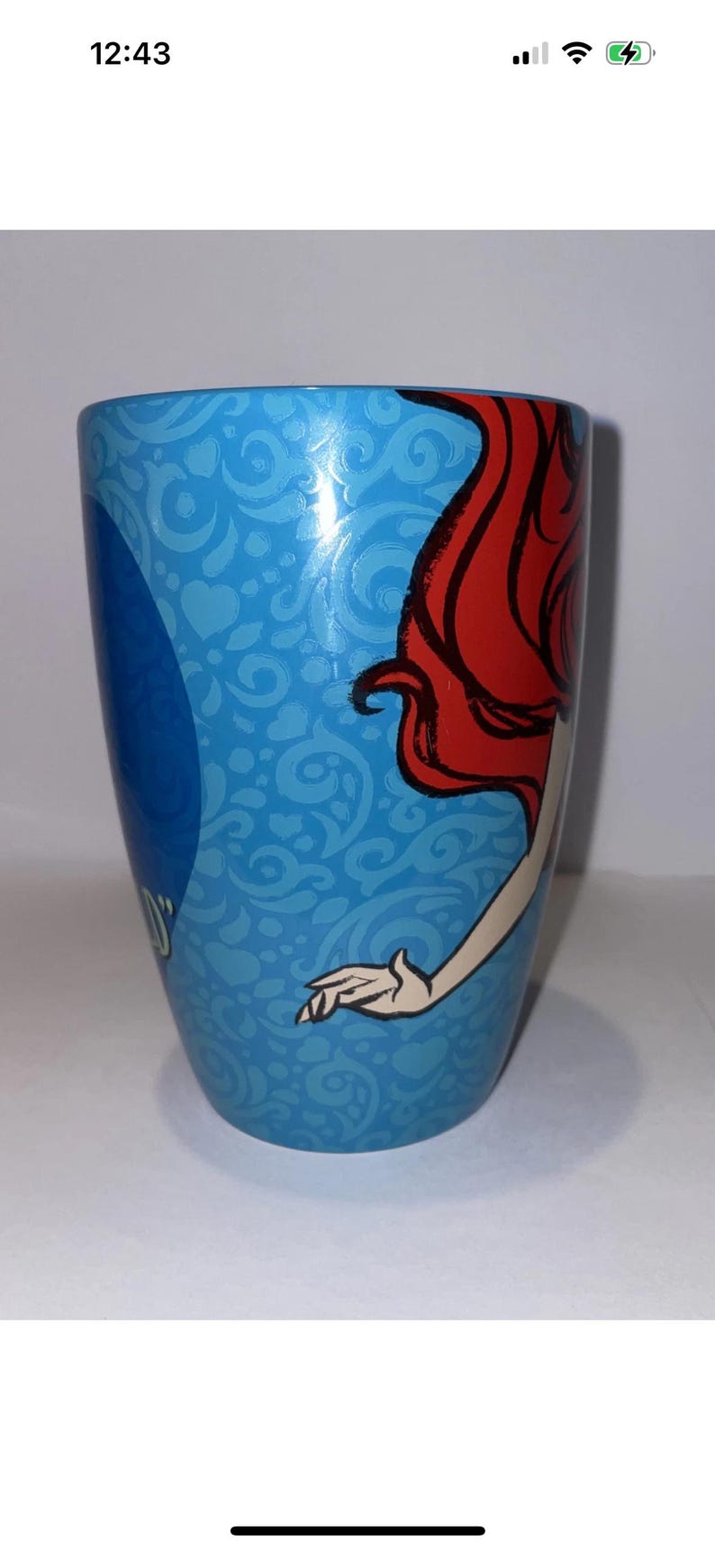 May include: A blue ceramic mug featuring a design of a character with red hair and a white arm. The mug has a light blue background with a pattern of swirls and hearts. The mug is cylindrical.