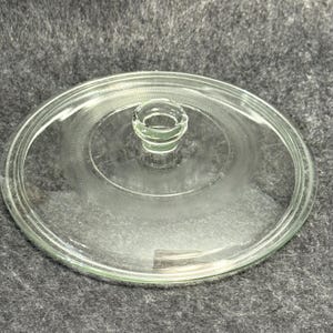 May include: A circular, clear glass lid featuring a central knob handle. The lid has a smooth, flat surface with a subtle, concentric textured ring pattern etched into the glass, providing a decorative detail around the center knob.