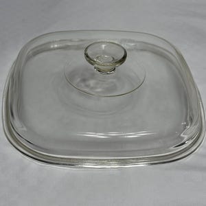 May include: A square, clear glass lid with rounded corners and a centered, circular knob handle. The lid features a slightly raised, flat top surface and a smooth, transparent finish, designed to fit a square cooking vessel or container.