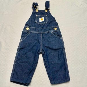 May include: Dark blue denim overalls featuring gold-tone metal buttons on the shoulder straps and waist. A square chest pocket displays a white Carhartt brand patch with a yellow logo. The garment includes contrast stitching and snap closures.