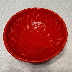 May include: A vibrant red ceramic bowl with a floral design embossed on the interior. The bowl has a glossy finish and a slightly irregular, organic shape. The bowl is approximately 8 inches in diameter, perfect for serving or display.