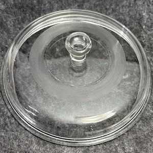 May include: A circular clear glass lid featuring a central knob handle and a decorative etched band with a fine cross-hatched texture. The lid has a raised outer rim and a smooth, transparent surface, designed to fit a container or dish.