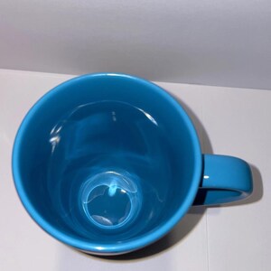 May include: A bright teal ceramic mug with a rounded body and a matching handle. The mug is empty and sits on a white surface. The mug is a solid colour with a glossy finish.