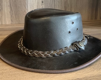May include: A brown leather cowboy hat with a braided leather band and a metal buckle. The hat has a wide brim and a tall crown.
