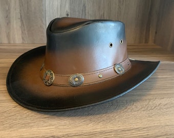 May include: A brown leather cowboy hat with a black brim and crown. The hat features decorative metal conchos around the brim and three eyelets on each side. This western-style hat is a classic accessory.