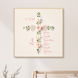 Puede incluir: Una cruz floral con las palabras "I AM" y "Eva" en ella. La cruz está rodeada de flores rosas y blancas. Las palabras "Strong, Capable, Never Alone, Chosen, Enough, Victorious" están escritas en la cruz.