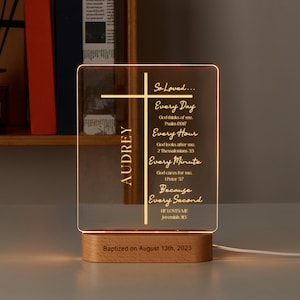 Puede incluir: Una base de madera con un panel de acrílico transparente que presenta una cruz y el nombre Audrey. El panel está iluminado con una luz blanca cálida y muestra un mensaje sobre el amor de Dios. El texto dice "So Loved... Every Day God thinks of me. Psalm 139:17 Every Hour God looks after me. 2 Thessalonians 3:3 Every Minute God cares for me. 1 Peter 5:7 Because Every Second HE LOVES ME Jeremiah 31:3". La base tiene la inscripción "Baptized on August 13th, 2023".