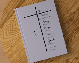 Op de afbeelding: Een lavendelkleurig notitieboek met een zwart kruis en de tekst "You are... Precious (1 Corinthians 6:20) Lovely (Daniel 12:3) Unique (Psalm 139:13) Forgiven (Psalm 103:12) Special (Ephesians 2:10) Healed (Isaiah 53:5)" en de naam "MIA" op de omslag.