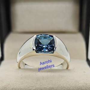 Op de afbeelding: Een zilveren ring met een grote, vierkante, blauwe edelsteen. De ring ligt in een zwarte doos met een witte binnenkant.