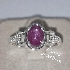 Op de afbeelding: Een zilveren ring met een roze ovale robijn, omringd door kleinere witte edelstenen. De ring ligt op een witte achtergrond met de tekst "Aarohi Jewellers".