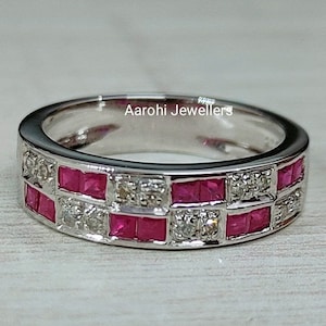 Puede incluir: Un anillo plateado con una banda que presenta filas alternas de piedras preciosas rosas brillantes cuadradas y pequeñas piedras preciosas transparentes. El anillo tiene un acabado pulido y un diseño moderno.