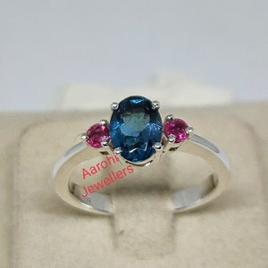 May include: A silver ring with a large blue oval gemstone and two smaller pink gemstones on either side. The ring is set in a white metal band. The text "Aaroh Jewellers" is visible on the band.