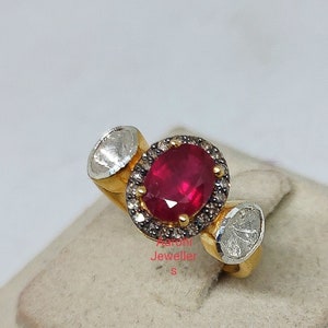 May include: A gold ring with a large oval ruby gemstone surrounded by smaller diamonds. The ring has two smaller diamond accents on either side of the ruby.