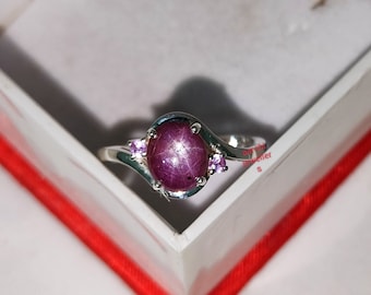 May include: A silver ring featuring a large oval ruby gemstone, surrounded by two smaller pink gemstones. The ring is displayed in a red velvet box.