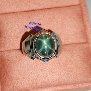 Puede incluir: Un anillo de plata con una piedra preciosa de zafiro estrella verde. El anillo tiene una banda texturizada y un engaste redondeado para la piedra. El texto "aarohi jewellers" es visible en la imagen.