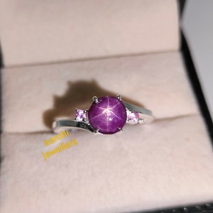 Op de afbeelding: Een zilveren ring met een grote paarse ster-saffier en twee kleinere roze saffieren. De ring ligt in een wit doosje met de tekst "aarohi jewellers" op de bodem.