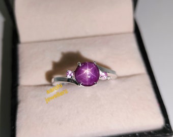 May include: A silver ring featuring a large purple star sapphire gemstone and two smaller pink sapphires. The ring is displayed in a white box with the inscription "aarohi jewellers" on the base.