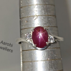May include: A silver ring featuring a red star ruby gemstone and two smaller white gemstones. The ring is displayed on a silver measuring tool.